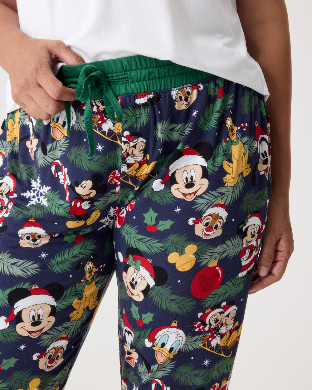 A close-up of a person wearing navy blue holiday-themed lounge pants with a green drawstring waist. The pants feature an all-over print of Disney characters like Mickey Mouse, Donald Duck, and Goofy wearing Santa hats, surrounded by green pine branches, red ornaments, and snowflakes.