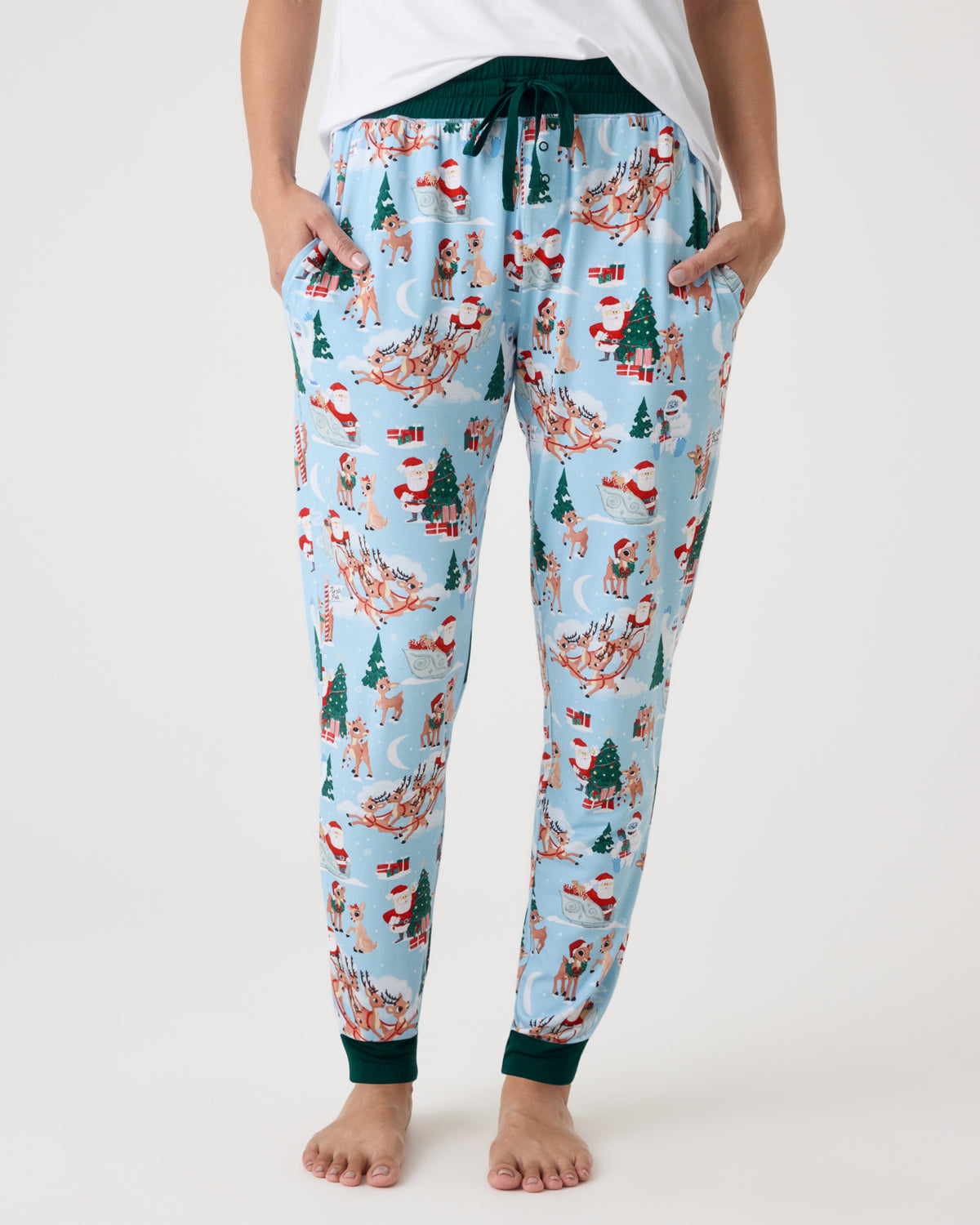 A person wearing light blue holiday-themed jogger pants with a festive print of Santa Claus, reindeer, Christmas trees, and sleighs, paired with a white top against a plain white studio background.