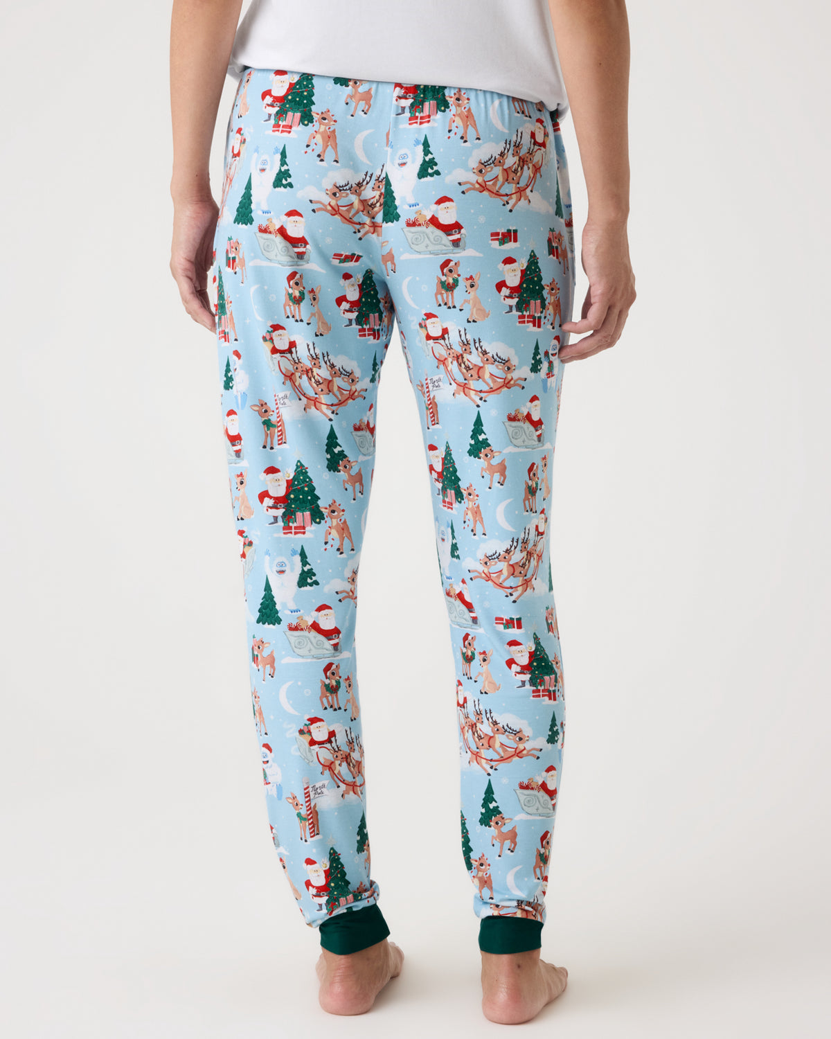 A person shown from the back wearing light blue pajama pants with an all-over Christmas print featuring Santa Claus, reindeer, trees, and sleighs, against a plain light background.