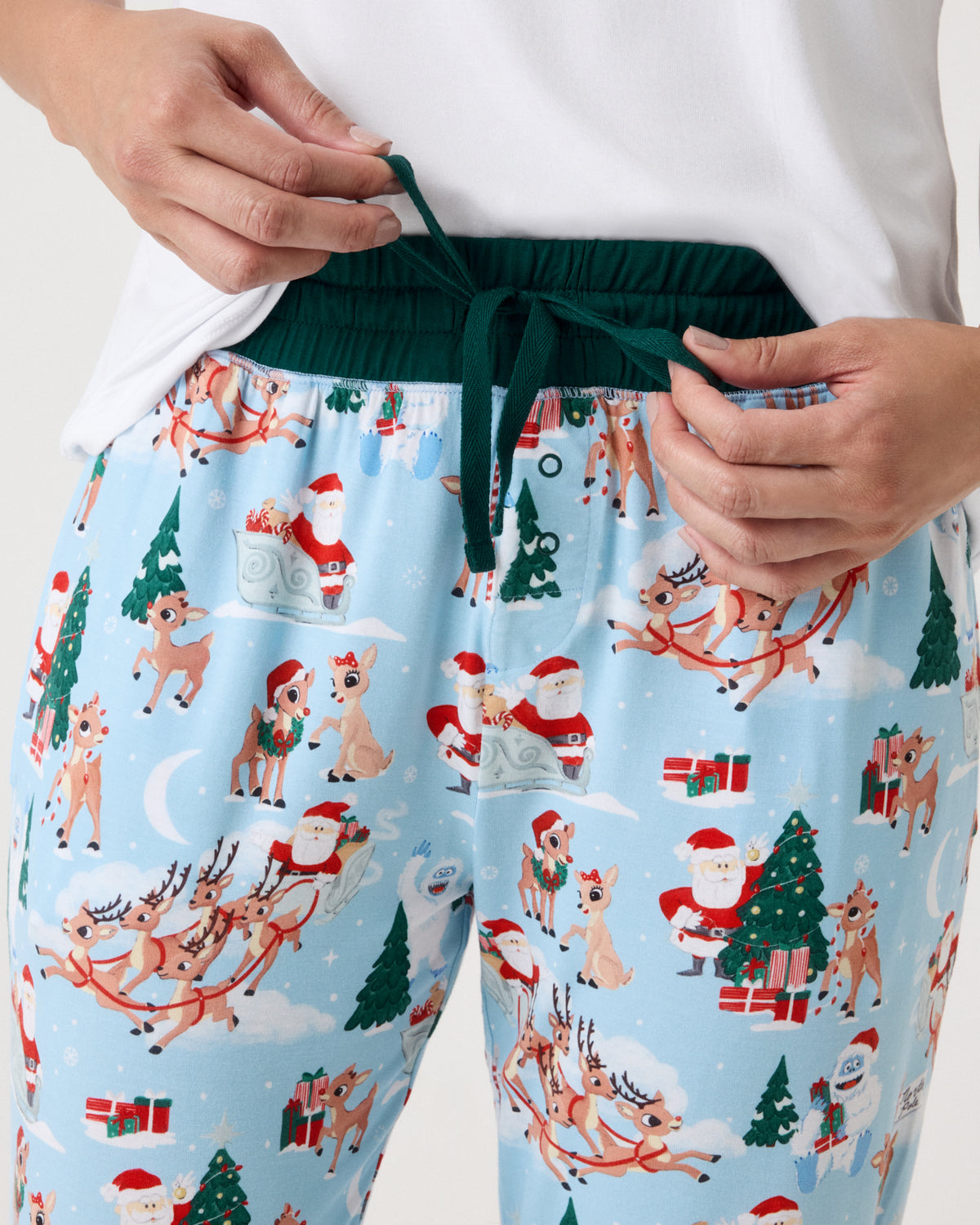 A close-up of a person wearing light blue Christmas-themed pajama pants with a festive print of Santa Claus, reindeer, dogs, and trees, paired with a white top and dark green drawstring waist.