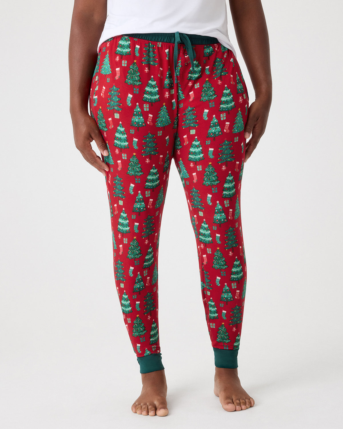 A person wearing red holiday pajama pants with a green Christmas tree and gift print, shown from the waist down against a light gray studio background.