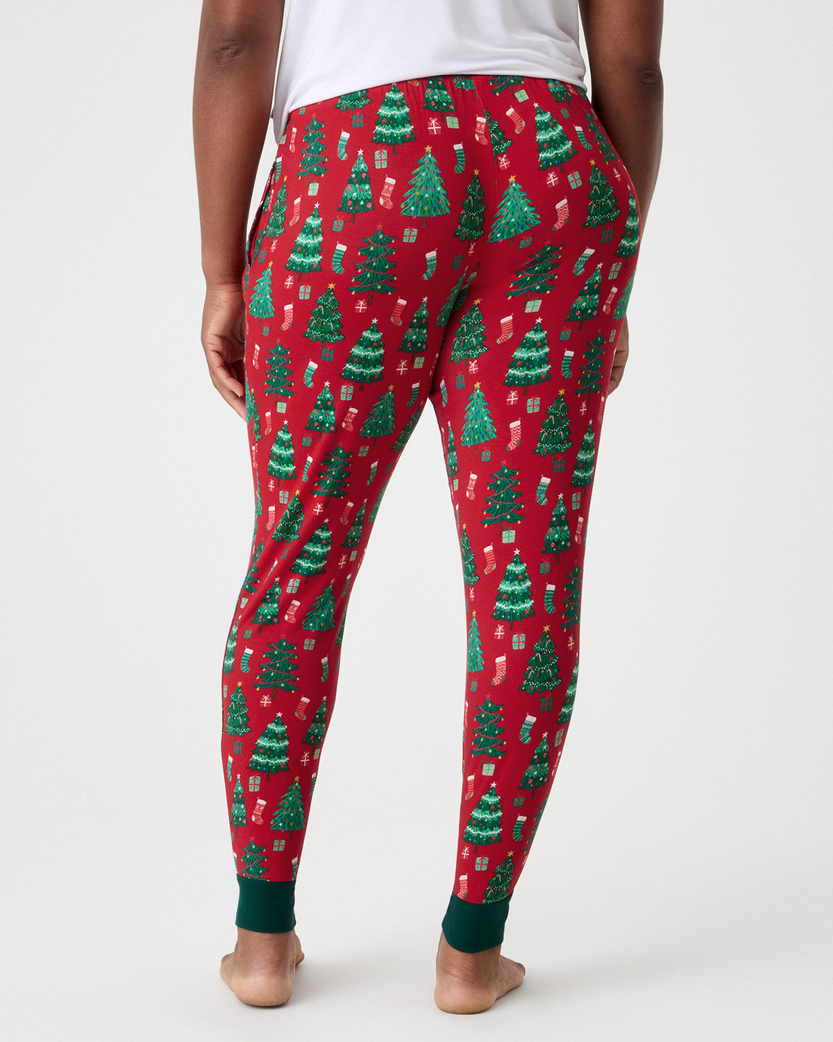 A person shown from the back wearing red holiday pants with a green Christmas tree and gift pattern, standing barefoot against a light gray studio background.
