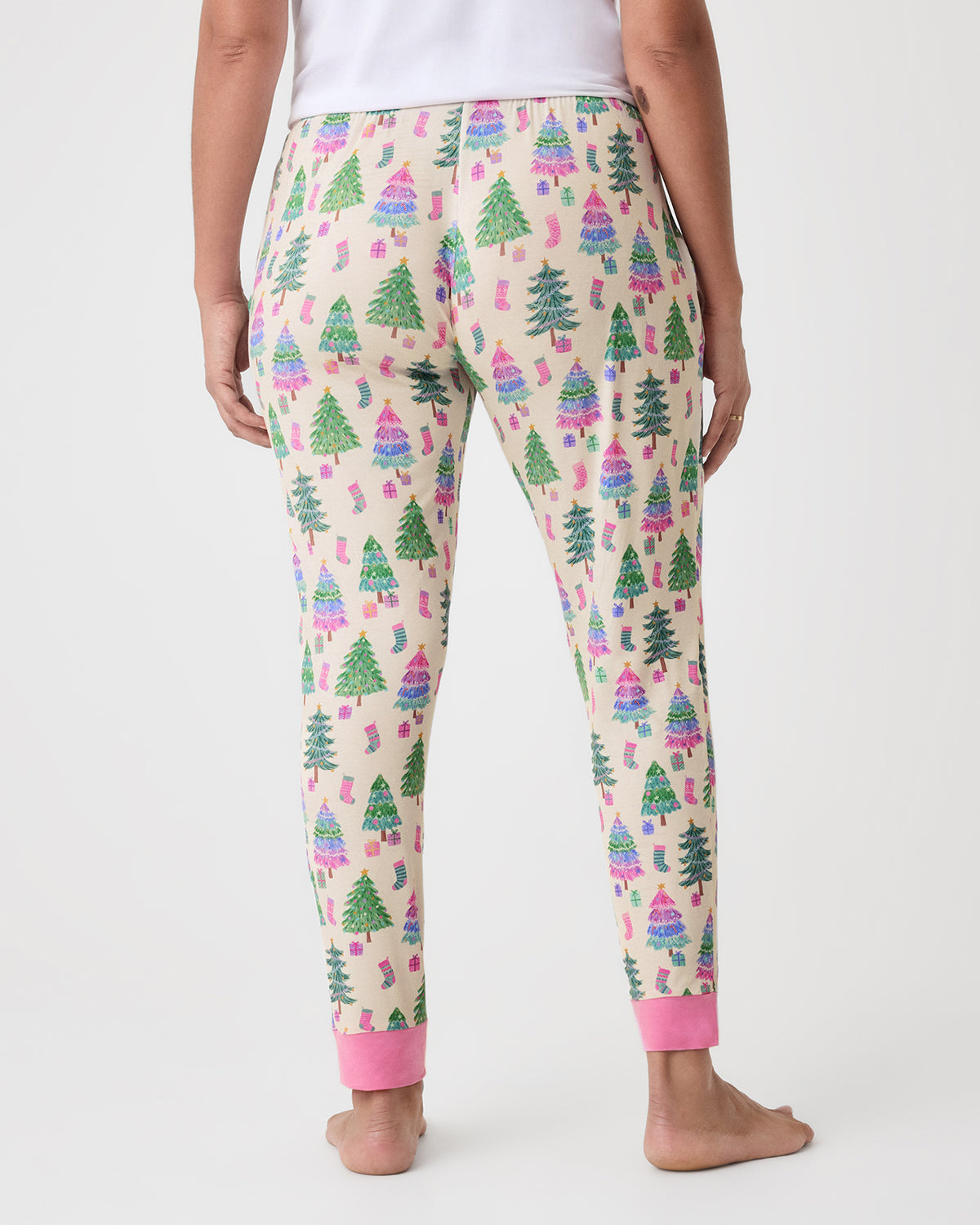A person shown from the back wearing cream pajama pants with an allover Christmas tree print in green, pink, and blue, with solid pink cuffs, standing barefoot against a light gray studio background.