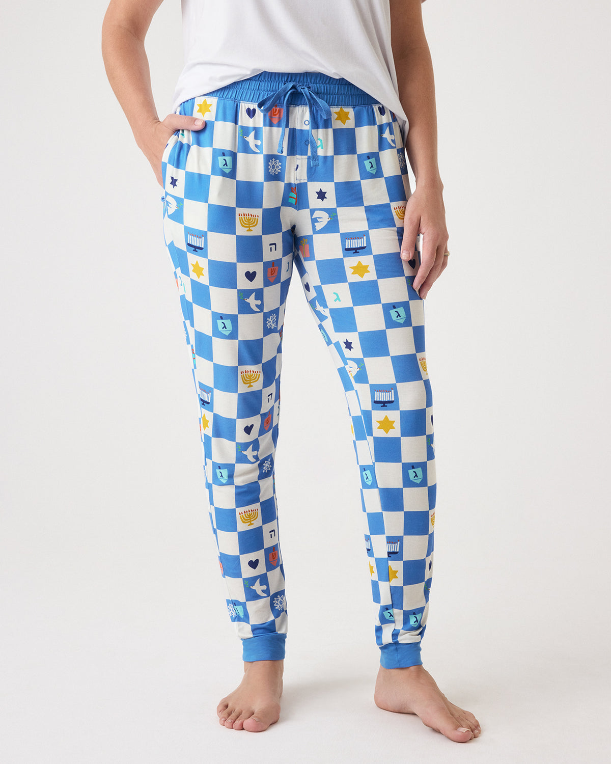 A person wearing blue and white checkered Hanukkah Checks Women's Pajama Pants with small multicolor printed squares, shown from the waist down against a light gray studio background.