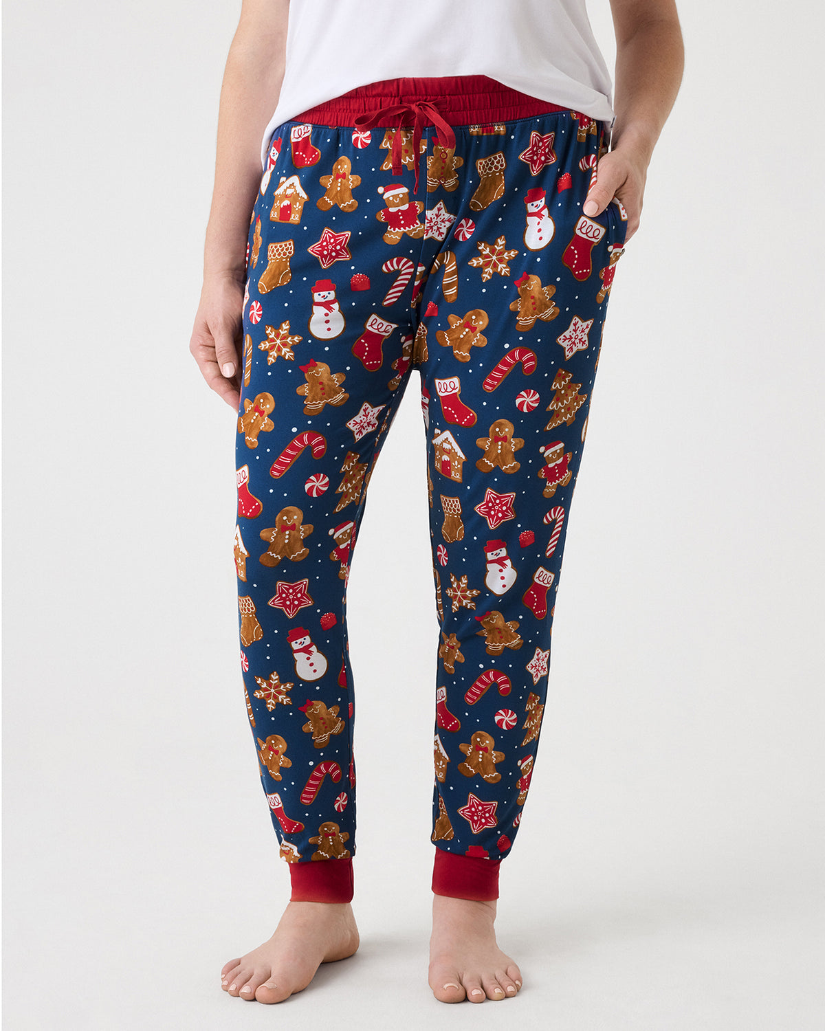 A person wearing navy blue pajama pants with a festive Christmas print of gingerbread men, snowmen, candy canes, and stockings, paired with a white top against a light gray studio background.
