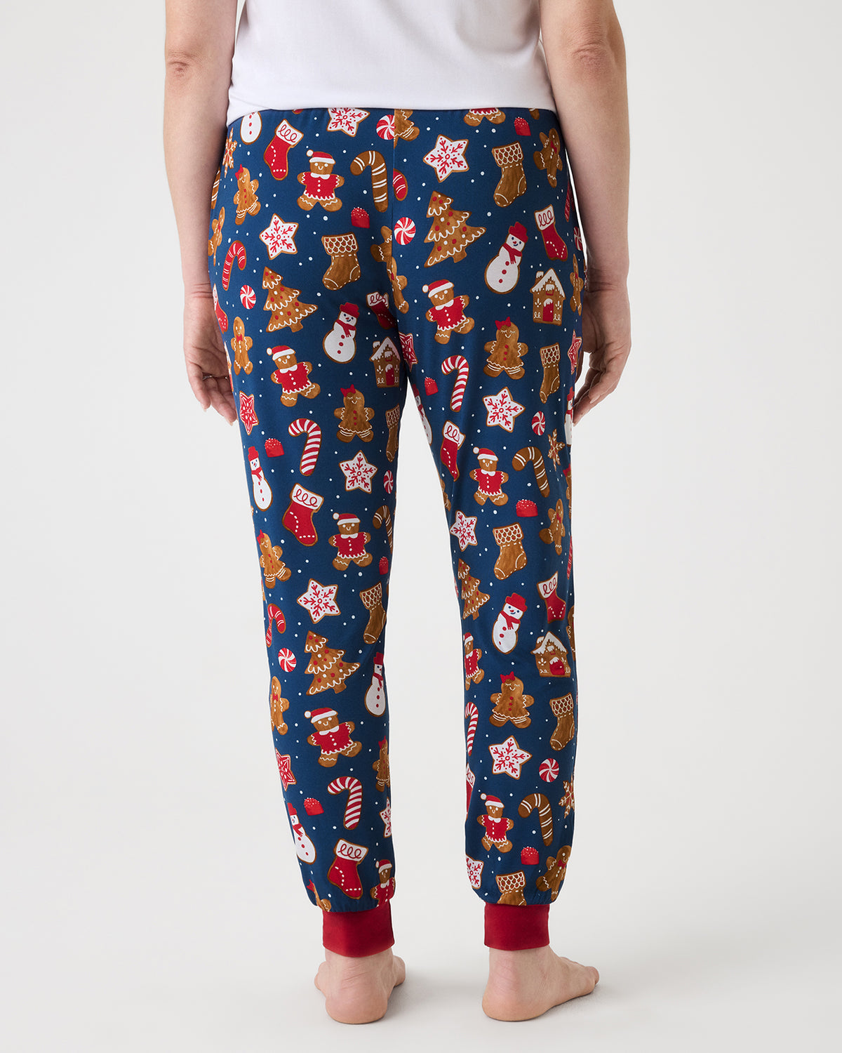 A person shown from the back wearing navy blue pajama pants covered in a festive Christmas print with gingerbread cookies, Santa Claus, candy canes, snowman, and stockings, against a light gray studio background.