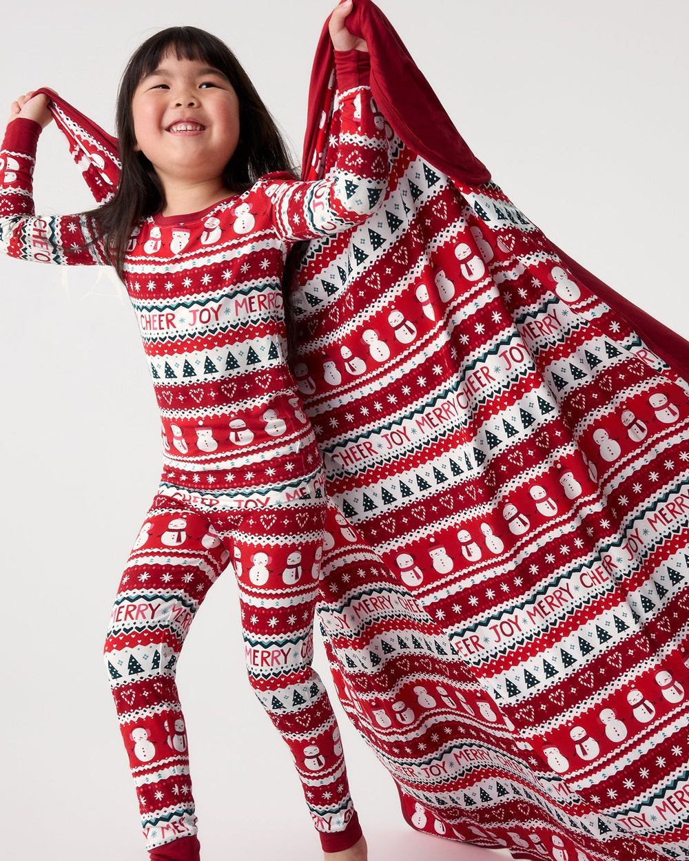A young girl smiling while wearing a red, white, and black Christmas-themed long-sleeve pajama set and holding up a matching patterned blanket against a plain light gray background.