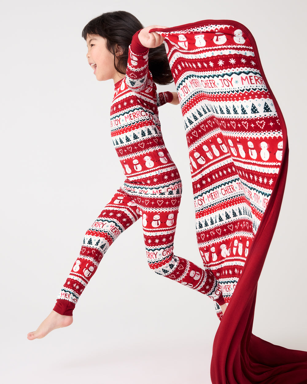 A child wearing a red, white, and black Christmas-themed knit one-piece jumpsuit and holding a matching knit blanket with holiday patterns, set against a plain light gray studio background.