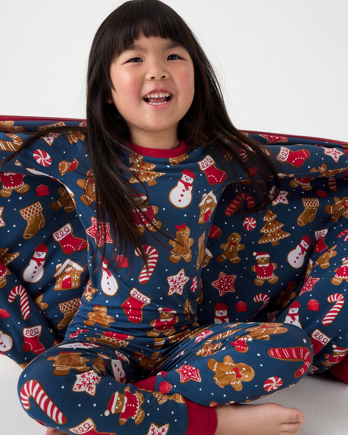 A smiling young girl wearing a navy blue Christmas-themed pajama set with a festive gingerbread, candy cane, snowman, and Santa pattern. She is posed against a clean white studio background.