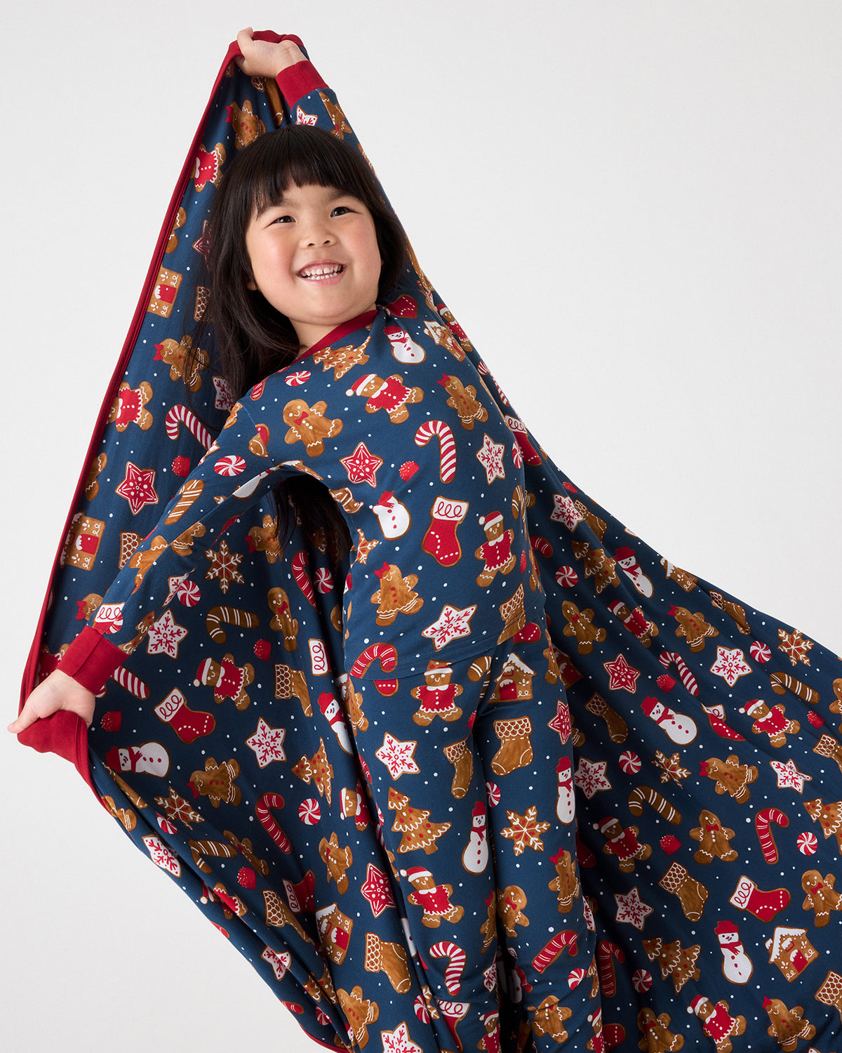 A smiling young girl wrapped in a navy blue holiday-themed blanket, holding up one corner to show the red lining. The blanket features a festive print of gingerbread cookies, Santa Claus, candy canes, snowmen, and stars in red, white, and brown, set against a solid light gray studio background.