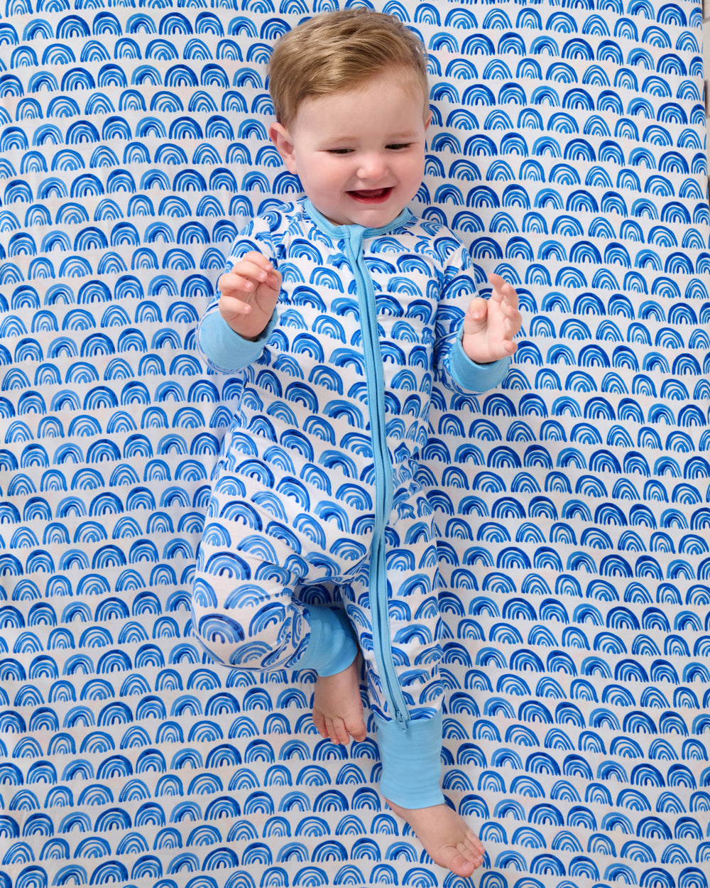 Baby laying on the Blue Rainbows Fitted Crib Sheet in a matching zippy