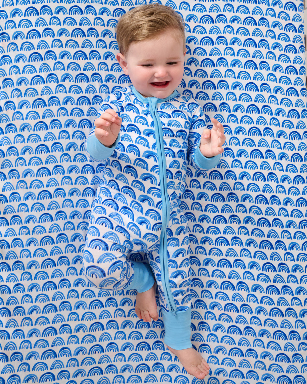 Baby laying on the Blue Rainbows Fitted Crib Sheet in a matching zippy