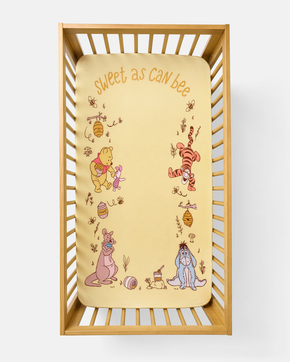 A baby crib mattress fitted sheet with a Winnie the Pooh theme, featuring illustrations of Pooh, Tigger, Eeyore, and Rabbit on a pale yellow background with the text 'sweet as can bee' at the top, shown inside a wooden crib.