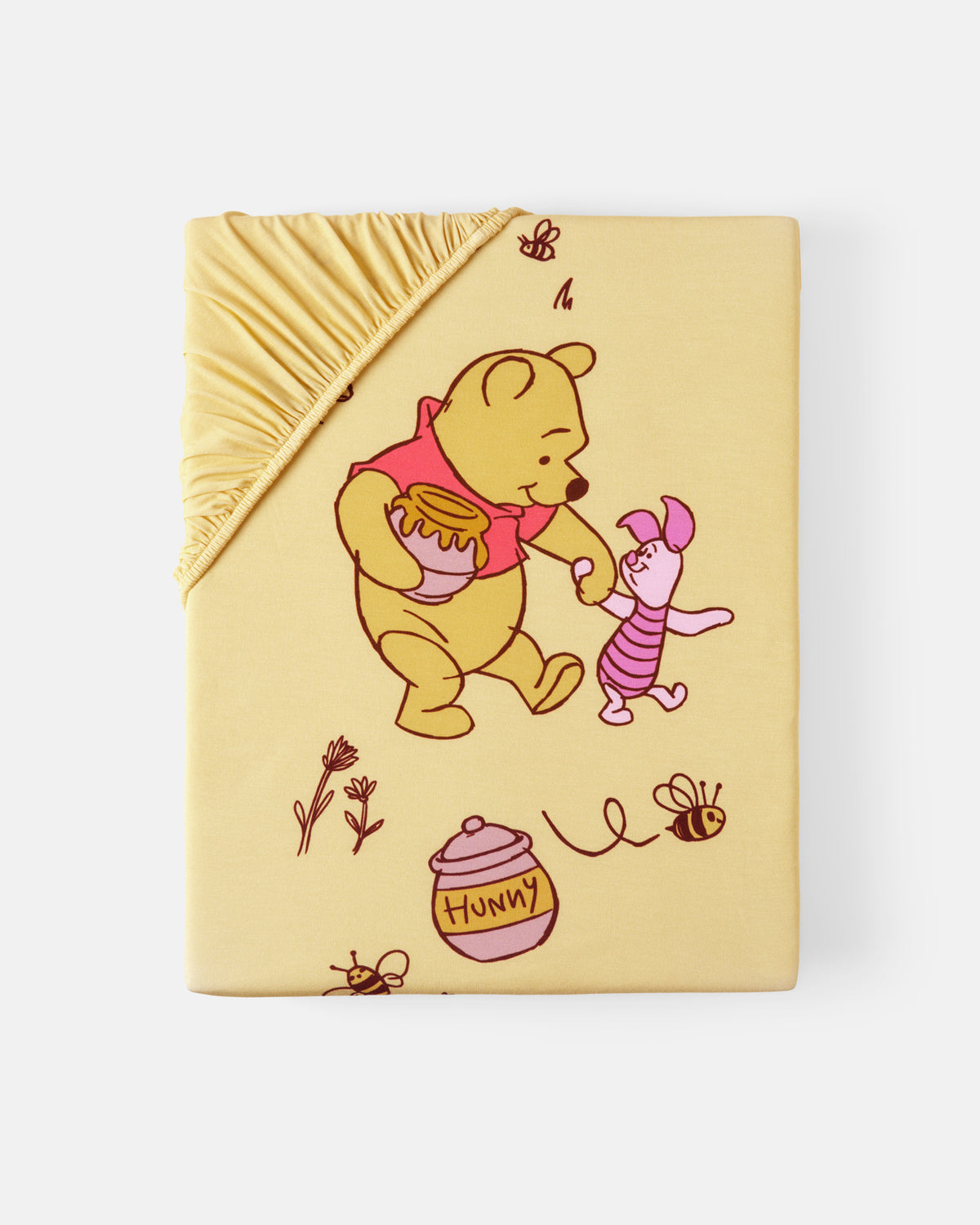 A pale yellow fitted sheet with a ruffled corner, printed with Winnie the Pooh and Piglet, honey pot, bees, and flowers on a light gray background.
