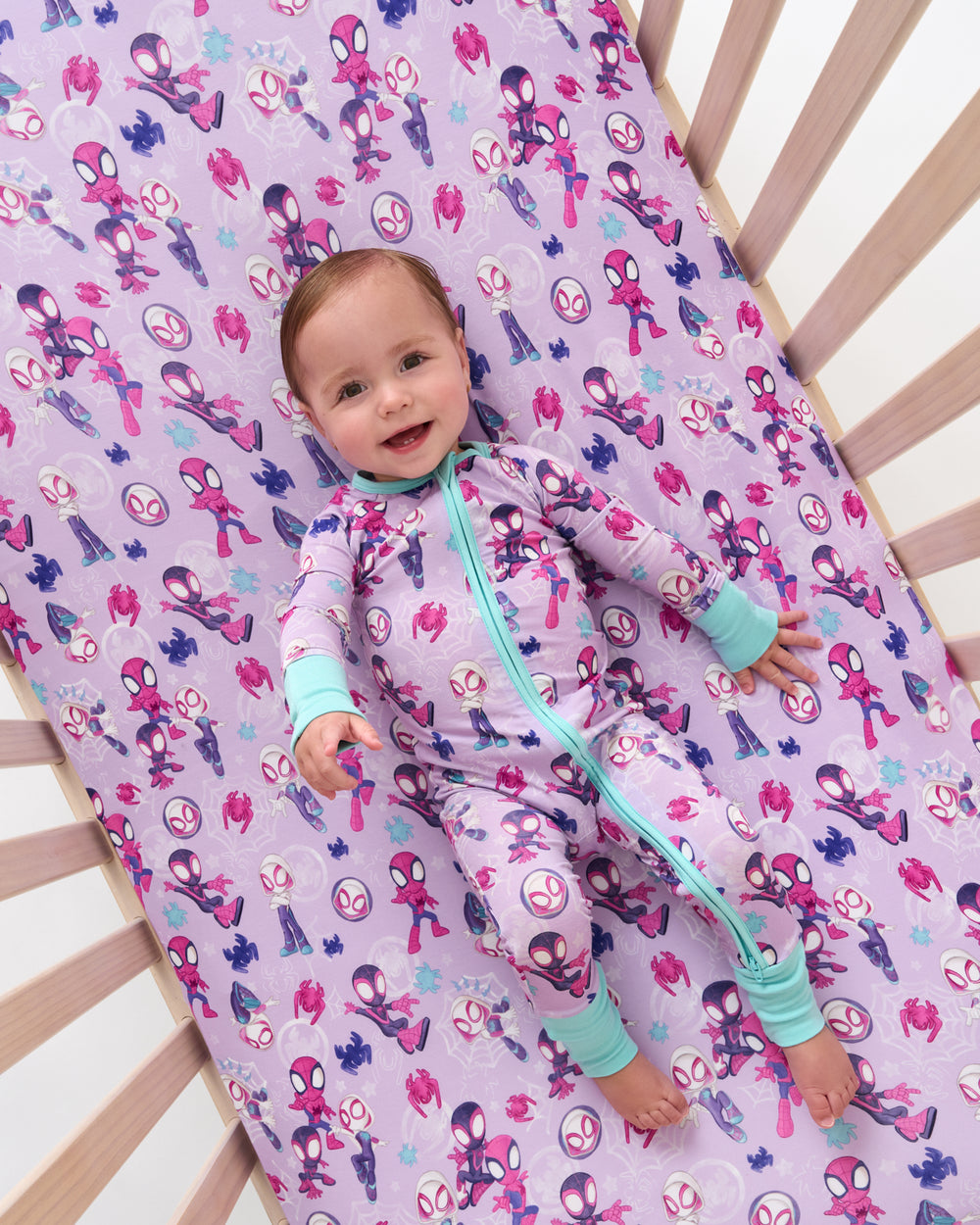 Aerial view of baby laying on the Purple Spidey & Friends Tiny Webs Fitted Crib Sheet in a matching footie pajama