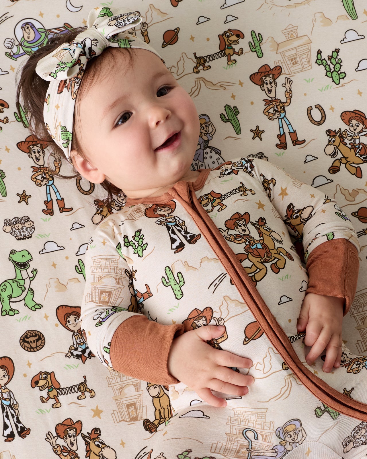 Baby wearing the Disney & Pixar Woody's Wild West Luxe Bow Headband and Zippy on a matching crib sheet