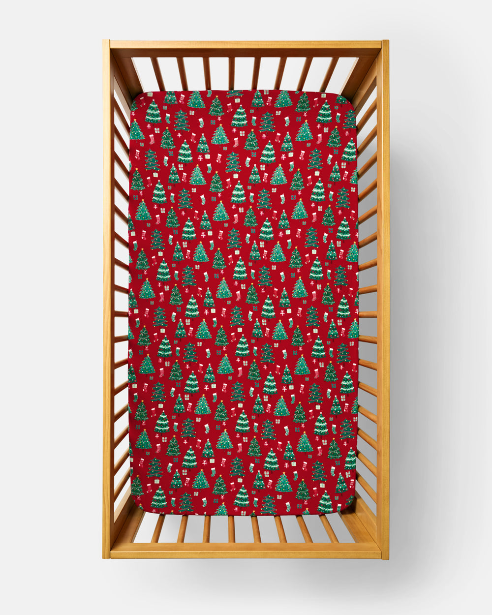 A red fitted crib sheet with a green Christmas tree pattern, shown inside a light wood crib on a white background.