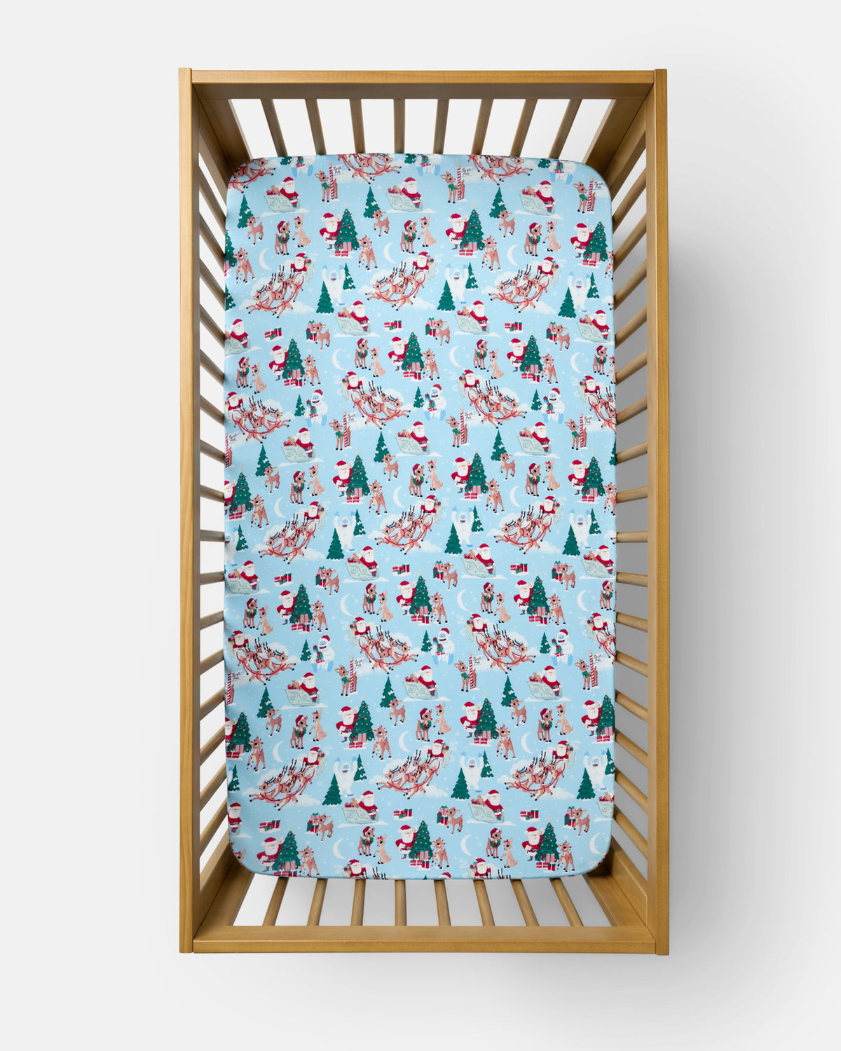 A light blue baby crib mattress cover with a festive Christmas print of Santa Claus, reindeer, and pine trees, shown inside a wooden baby crib.