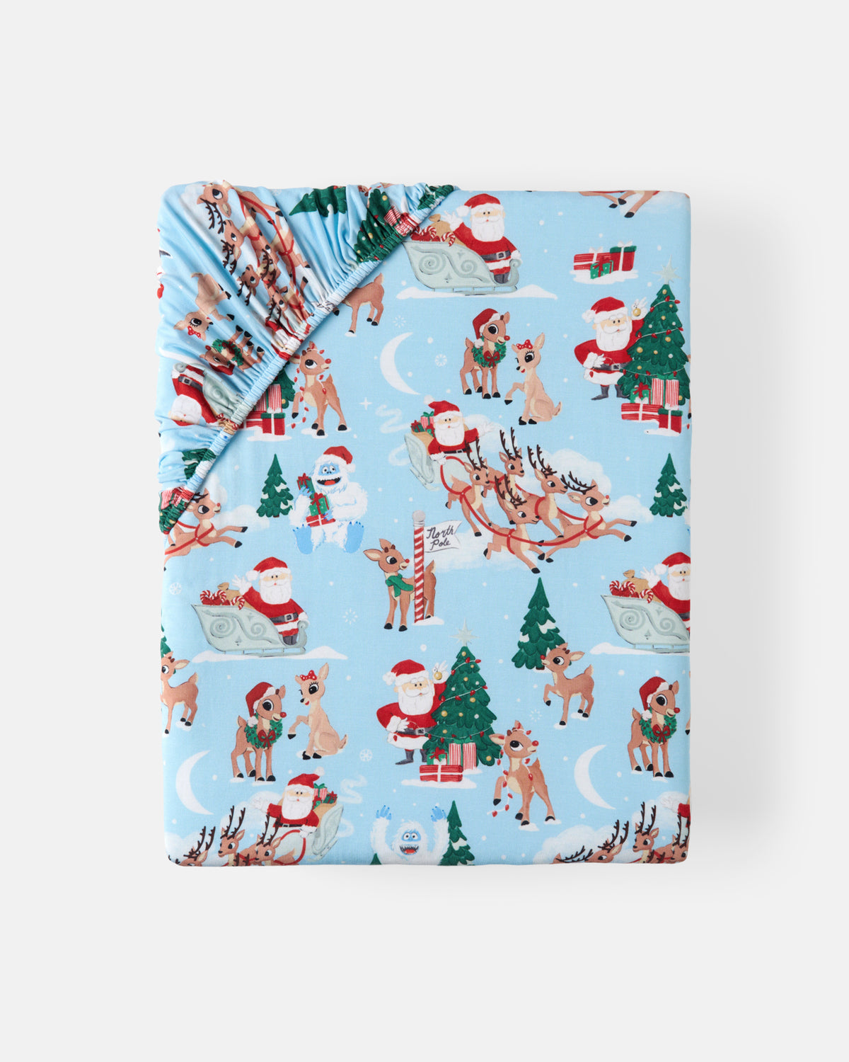 A folded baby sheet with a festive Christmas print, featuring Santa Claus, reindeer, Christmas trees, and snowflakes on a light blue background, shown against a plain white studio backdrop.