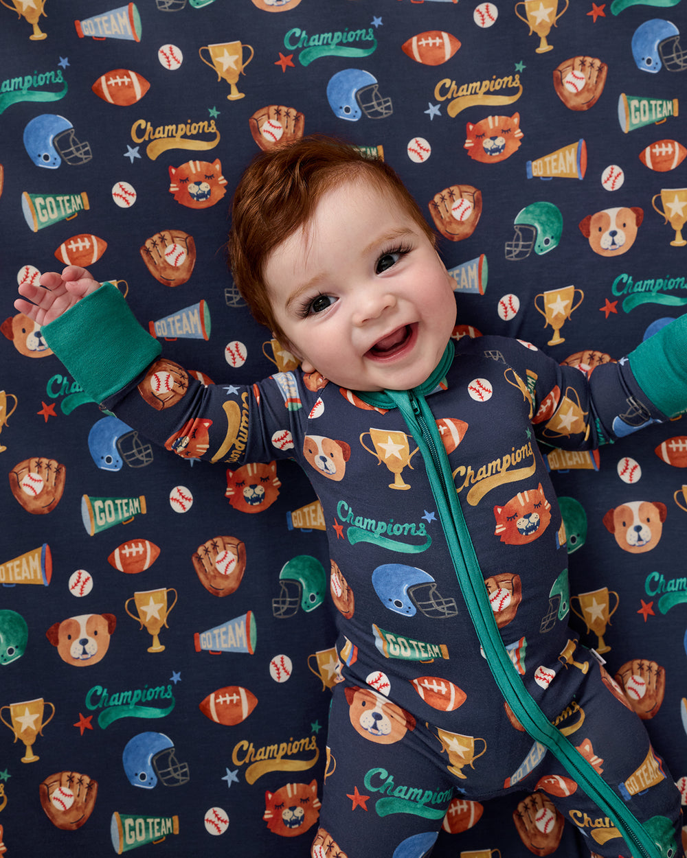 Baby laying on the Team Spirit Fitted Crib Sheet wearing a matching zippy