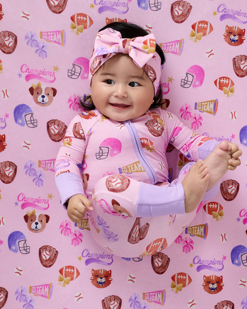 Baby laying on the Team Spirit Pink Fitted Crib Sheet wearing a matching footie pajama