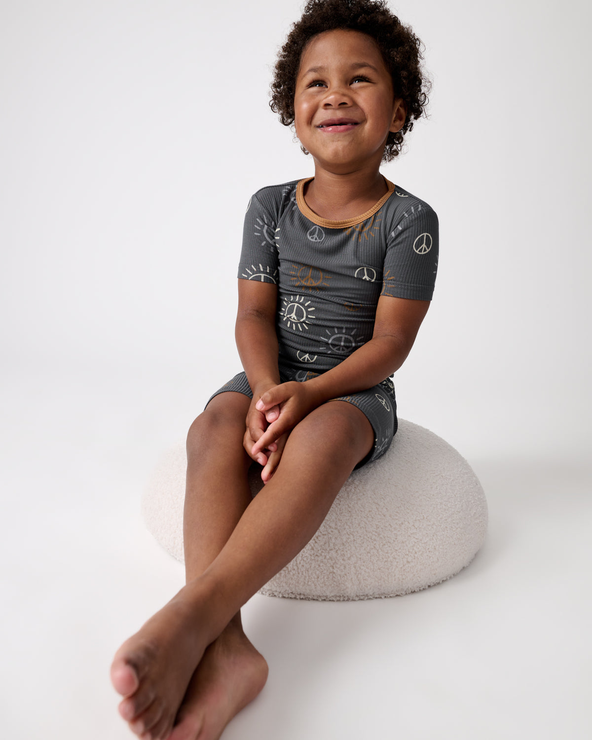 Boy sitting on a white ottoman wearing the Peace & Love Ribbed Two-Piece Short Sleeve & Shorts Pajama Set