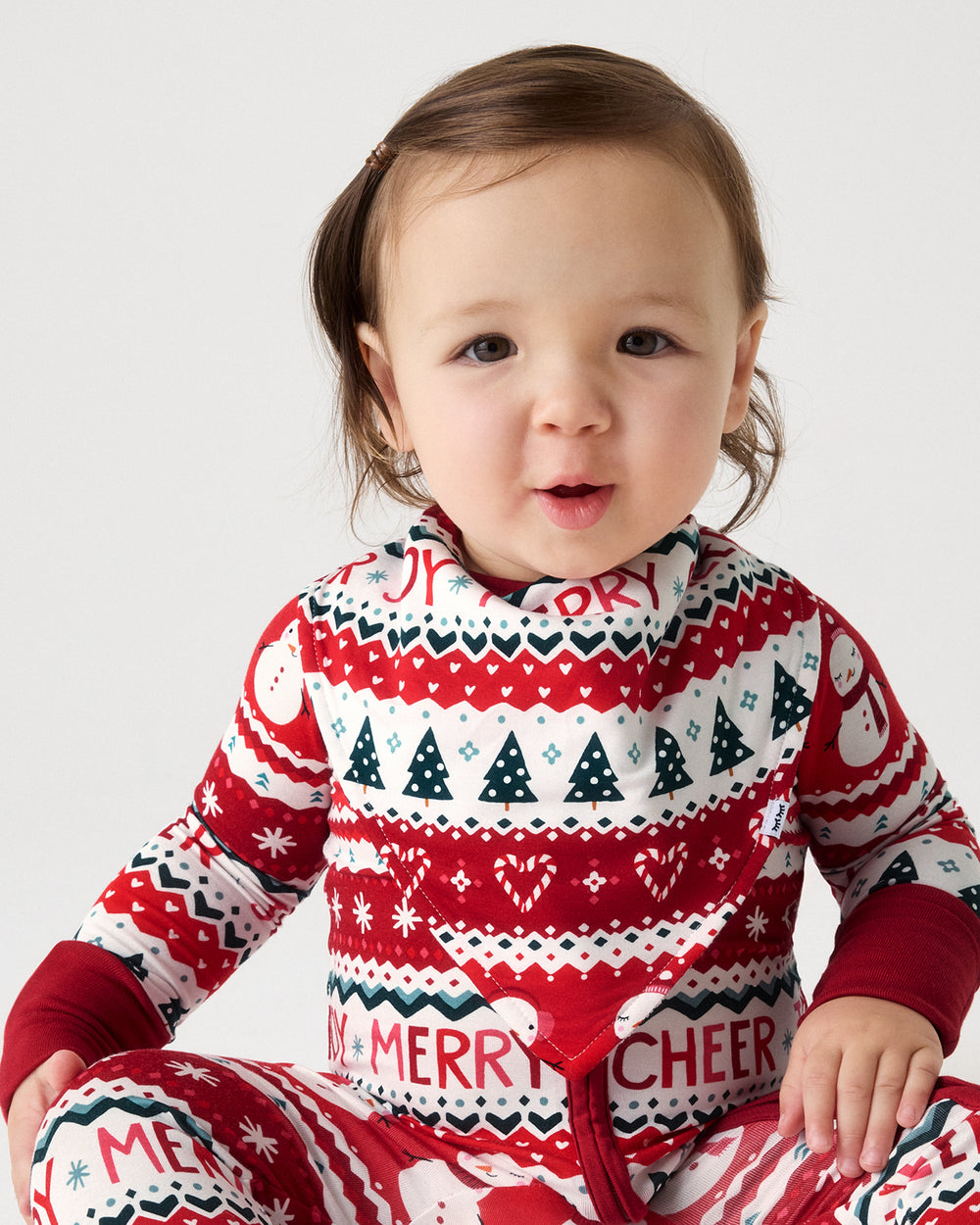 A baby wearing a festive red, white, and navy holiday-themed long-sleeve one-piece pajama with 'Merry Cheer' text, photographed against a light gray studio background.
