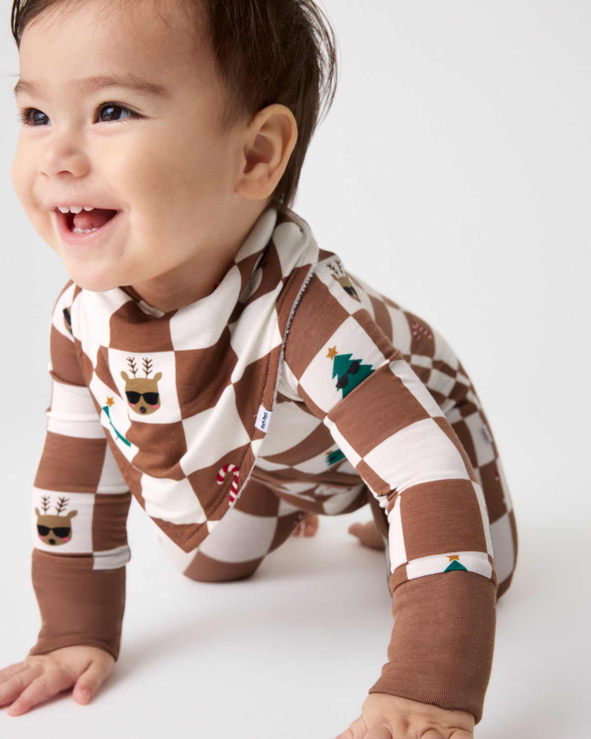 A baby crawling on a white studio floor wearing a brown and cream checkered long-sleeve bodysuit with holiday-themed printed squares.