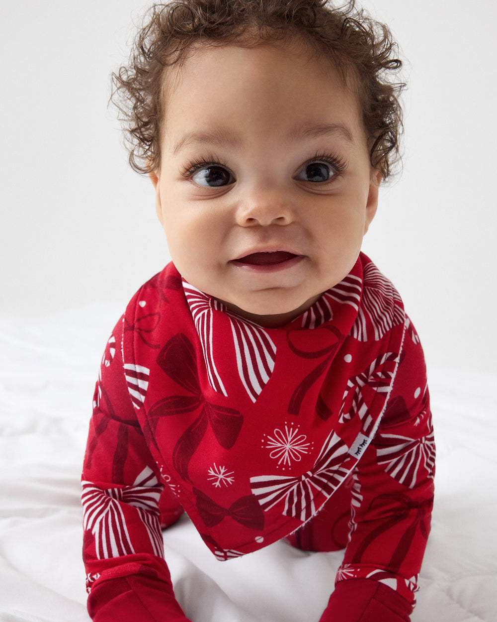 A baby wearing a red long-sleeve zipper PJ with a white and red bow pattern, photographed from the chest up against a plain light background.