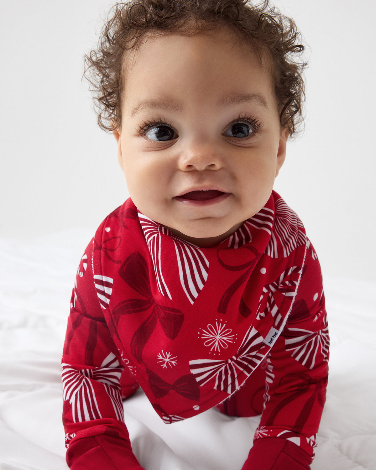 A baby wearing a red long-sleeve zipper PJ with a white and red bow pattern, photographed from the chest up against a plain light background.