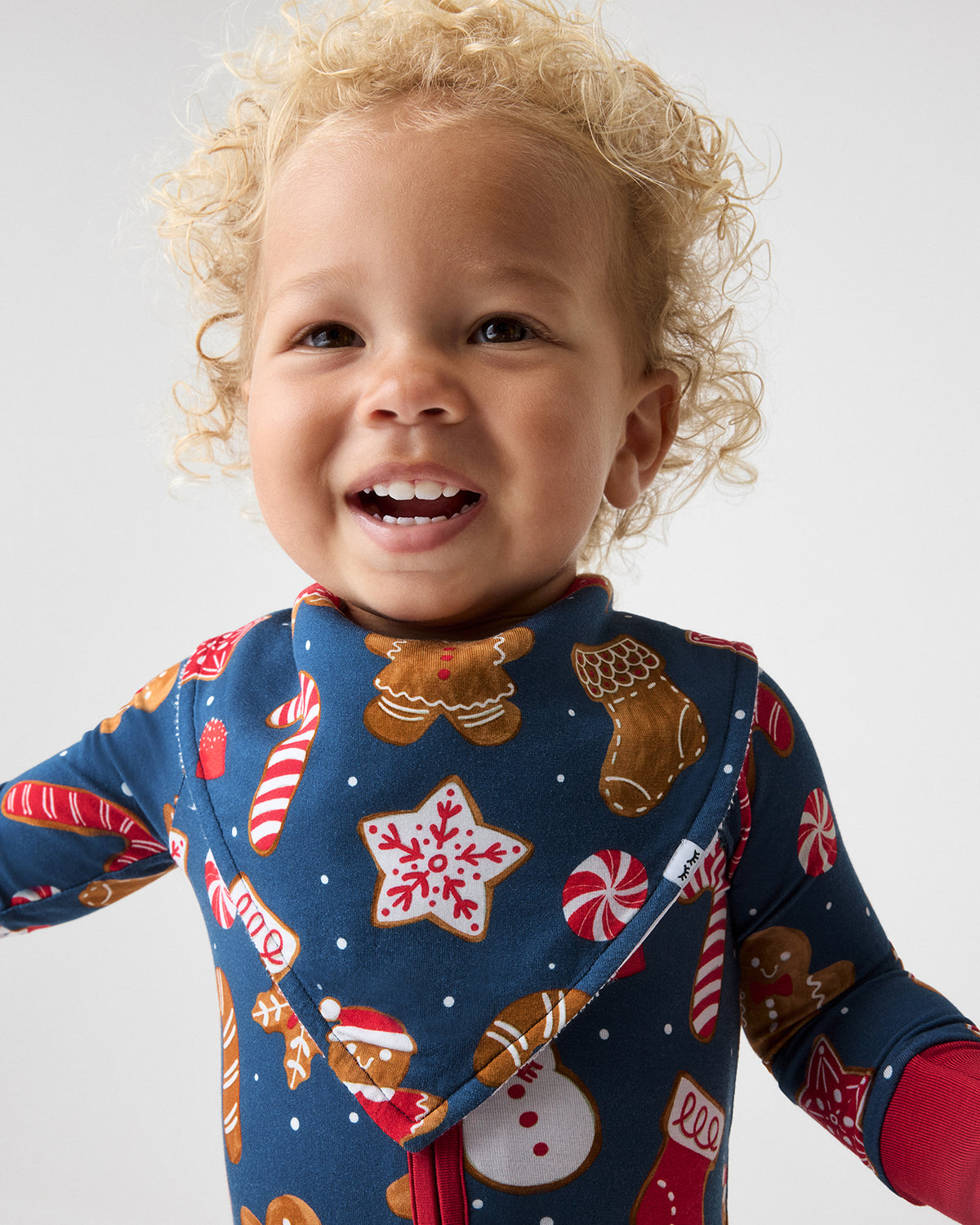A smiling toddler with curly blonde hair wearing a navy blue holiday-themed long-sleeve footie pajama with a front zipper and matching bib, printed with gingerbread cookies, candy canes, snowman, and stockings on a light gray studio background.