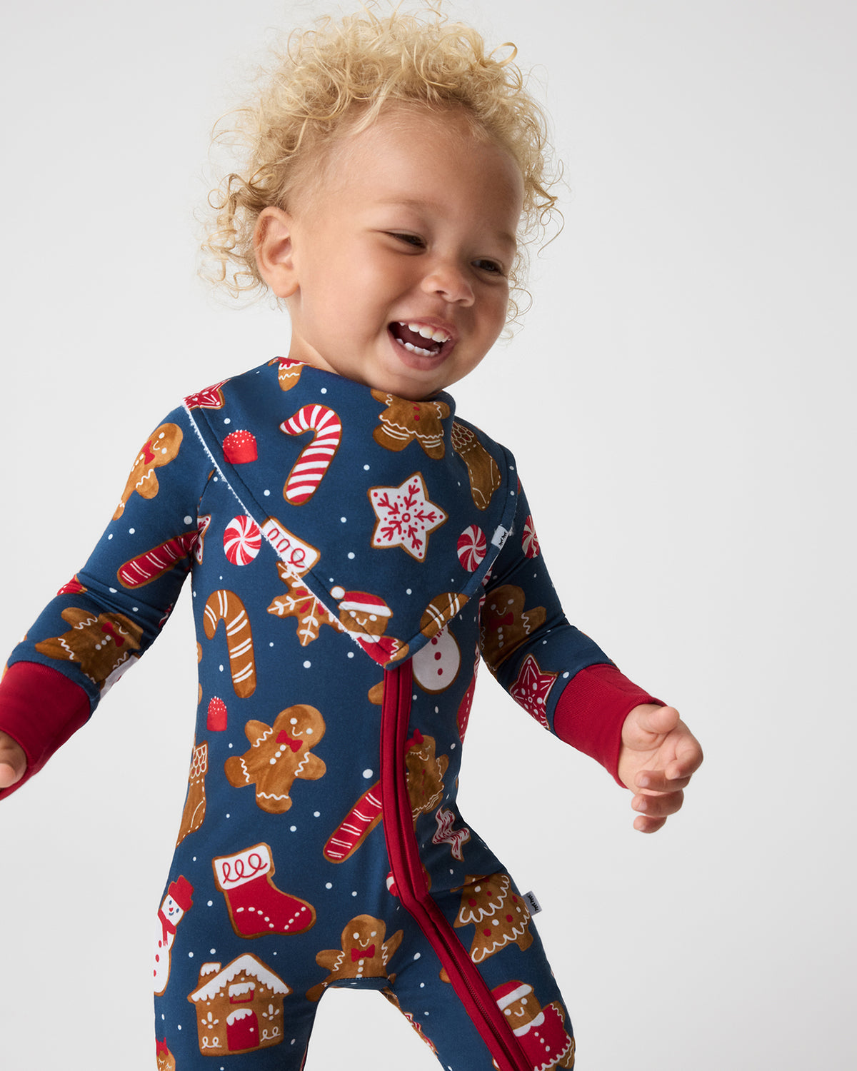 A toddler wearing a festive navy blue one-piece pajama and bib with gingerbread men, candy canes, and stars, smiling and posing against a light gray background.