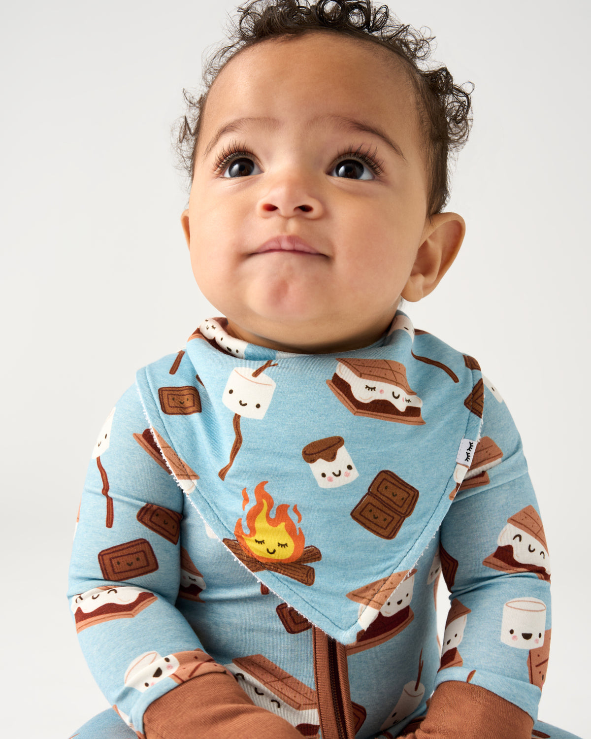 A baby wearing a light blue long-sleeve zip-up onesie with a s'mores print, including marshmallows, chocolate bars, and a campfire, against a plain light gray background.