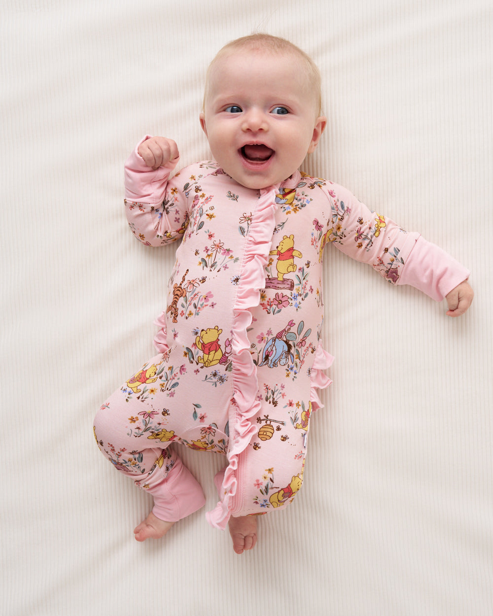 A baby lying on a cream-colored textured blanket, wearing a pink long-sleeve footless sleeper with ruffles and a colorful Winnie the Pooh print.