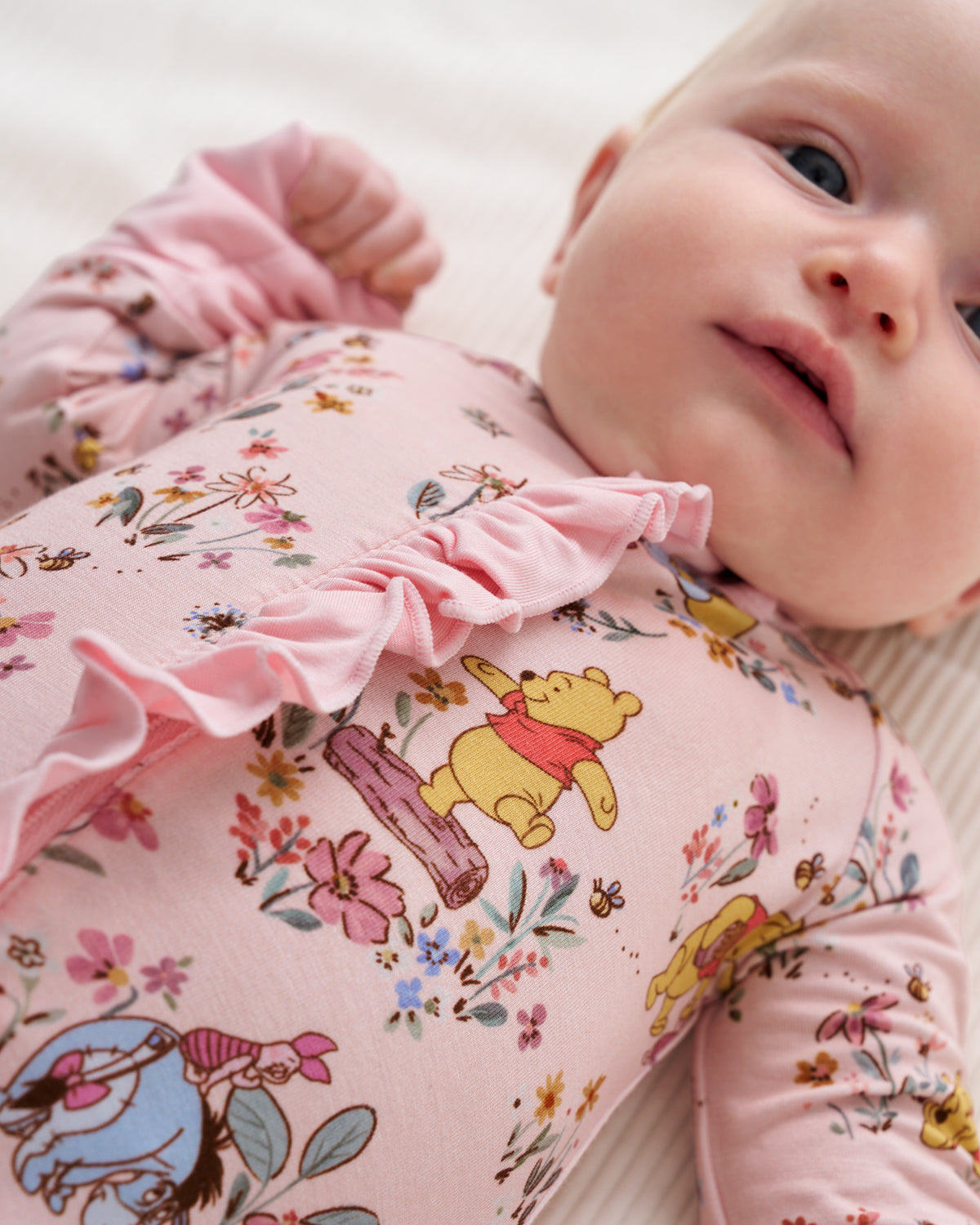 A close-up of a baby lying on a light cream surface, wearing a pink long-sleeve romper with a Winnie the Pooh floral print and a ruffled neckline.
