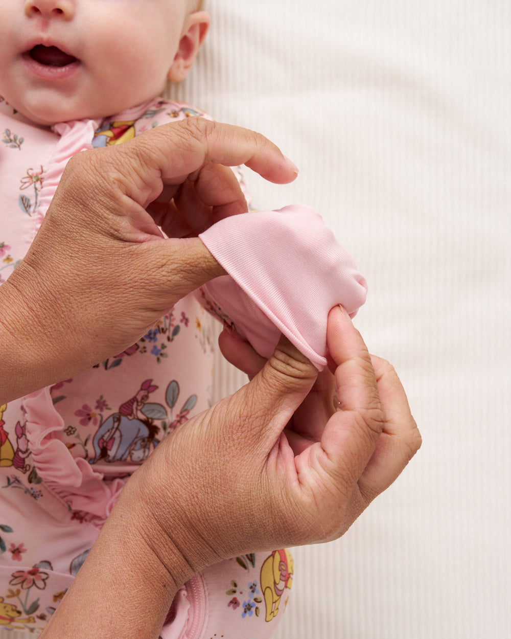 Close-up of an adult's hands fastening a pink fabric diaper cover or diaper on a baby wearing a pink floral onesie, set against a soft white textured background.