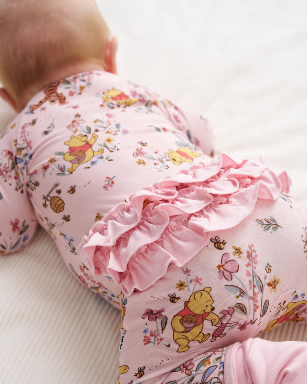 A baby lying on their stomach wearing a pink long-sleeve bodysuit with a Winnie the Pooh bear and floral print, featuring a large pink ruffle detail around the waist.
