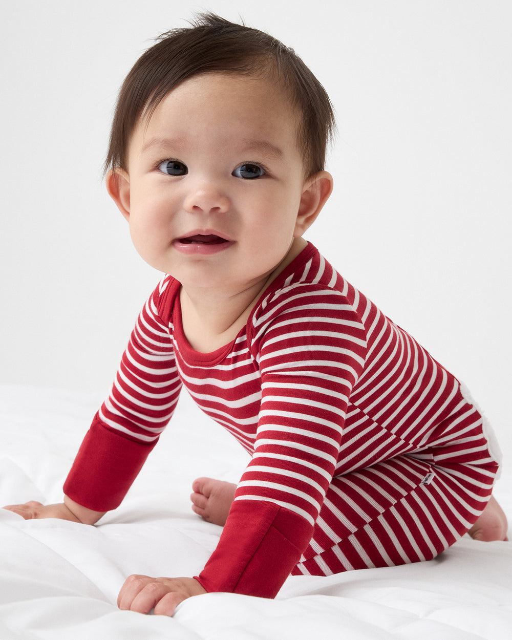 A baby wearing a red and white striped long-sleeve pajama top, crawling on a white sheet against a light studio background.