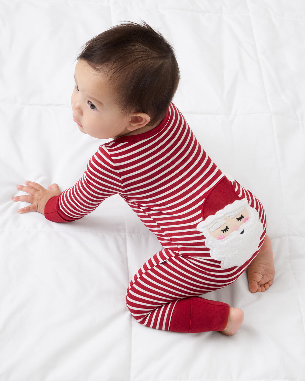 A baby wearing a red and white striped Christmas pajama with a Santa Claus face applique, crawling on a white blanket.