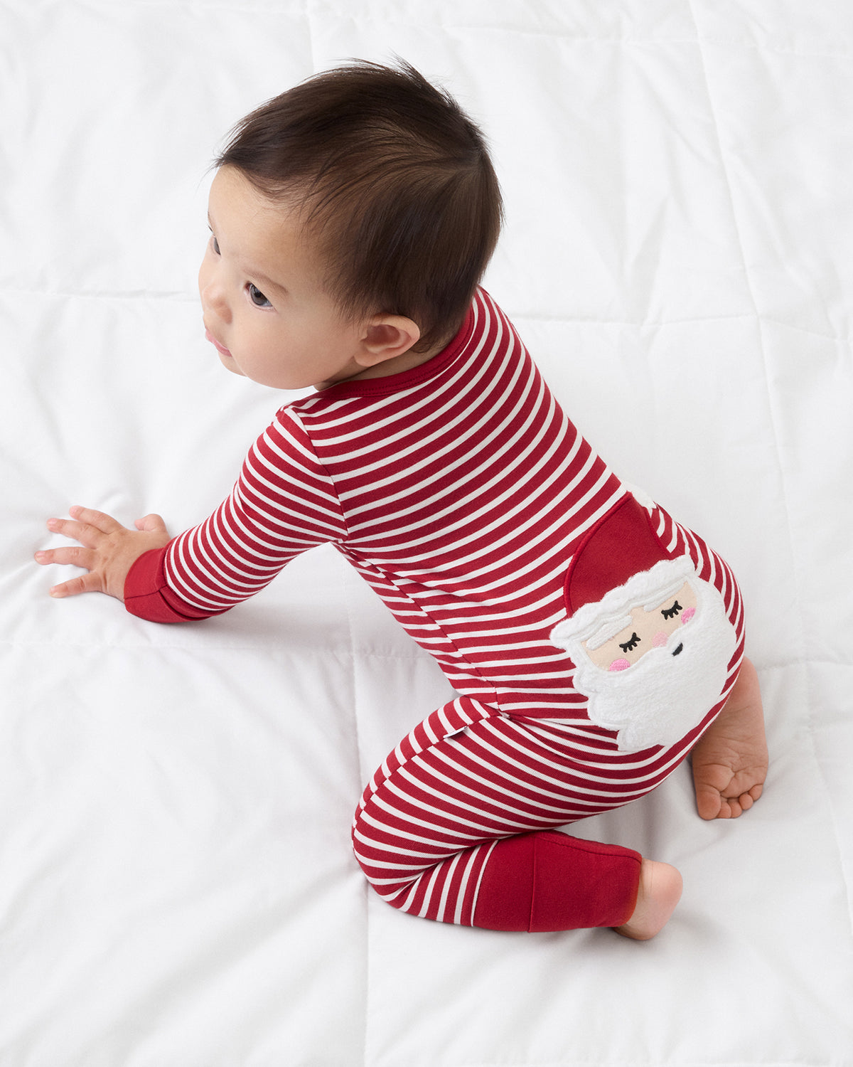A baby wearing a red and white striped Christmas pajama with a Santa Claus face applique, crawling on a white blanket.