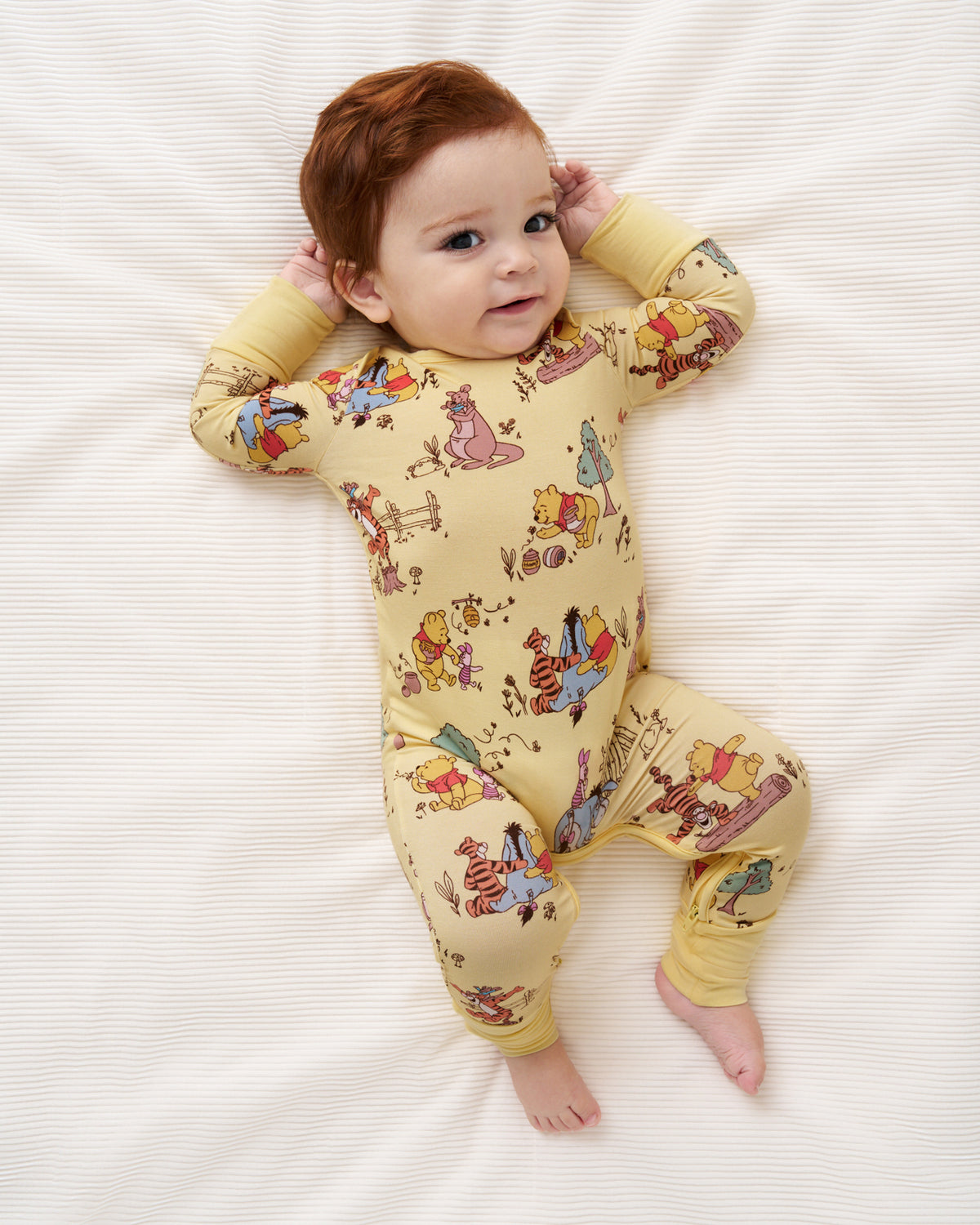 A baby lying on a white textured blanket, wearing a long-sleeve footed sleeper in a pale yellow color with a colorful cartoon print.
