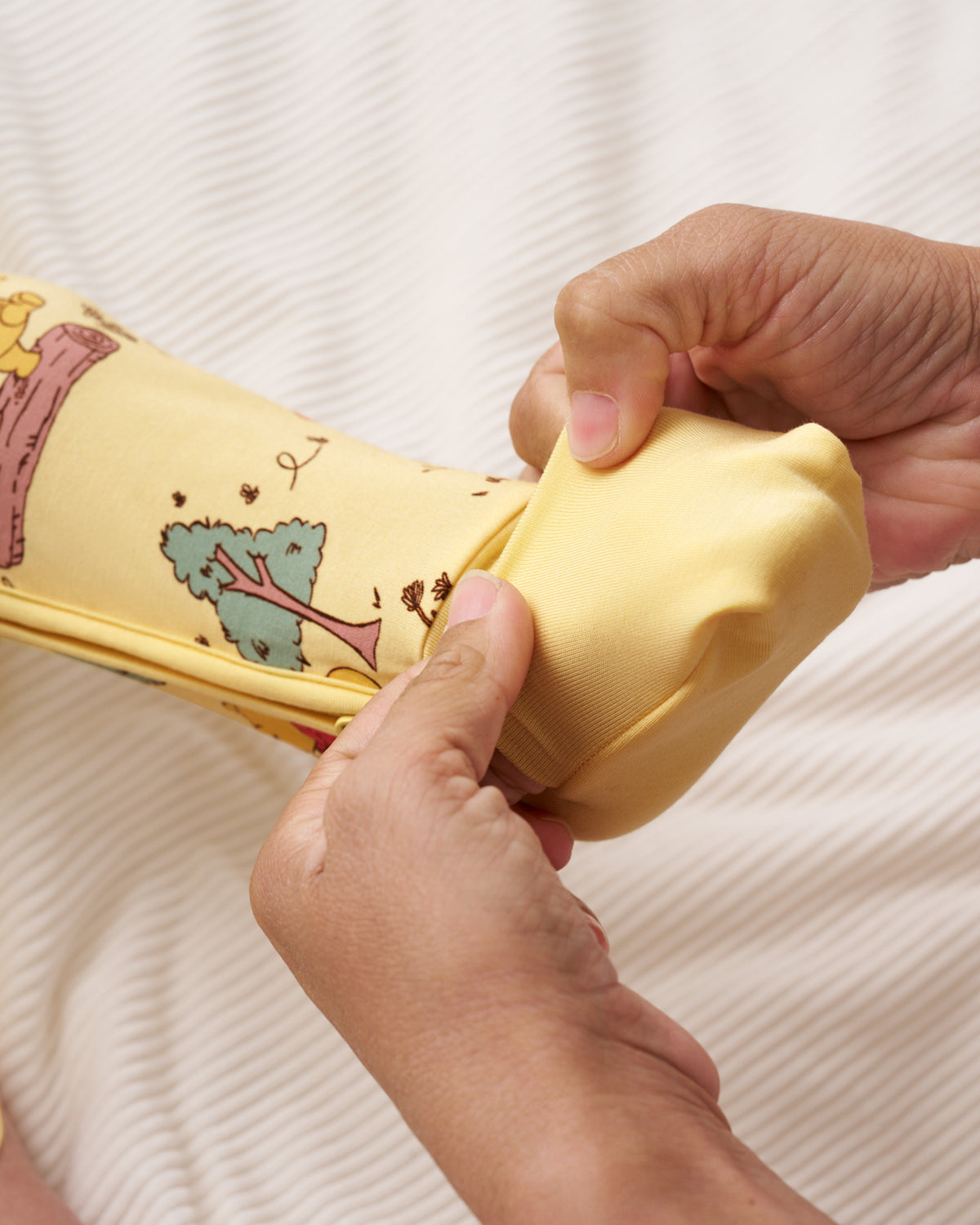 Close-up of hands folding a yellow satin-like fabric sleeve with a printed cartoon tree design, set against a ribbed cream fabric background.