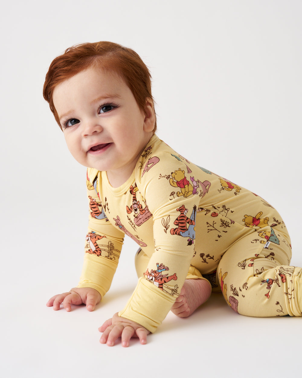 A baby with red hair crawling on a white studio floor, wearing a pale yellow long-sleeve footed romper covered in a colorful Winnie the Pooh print.
