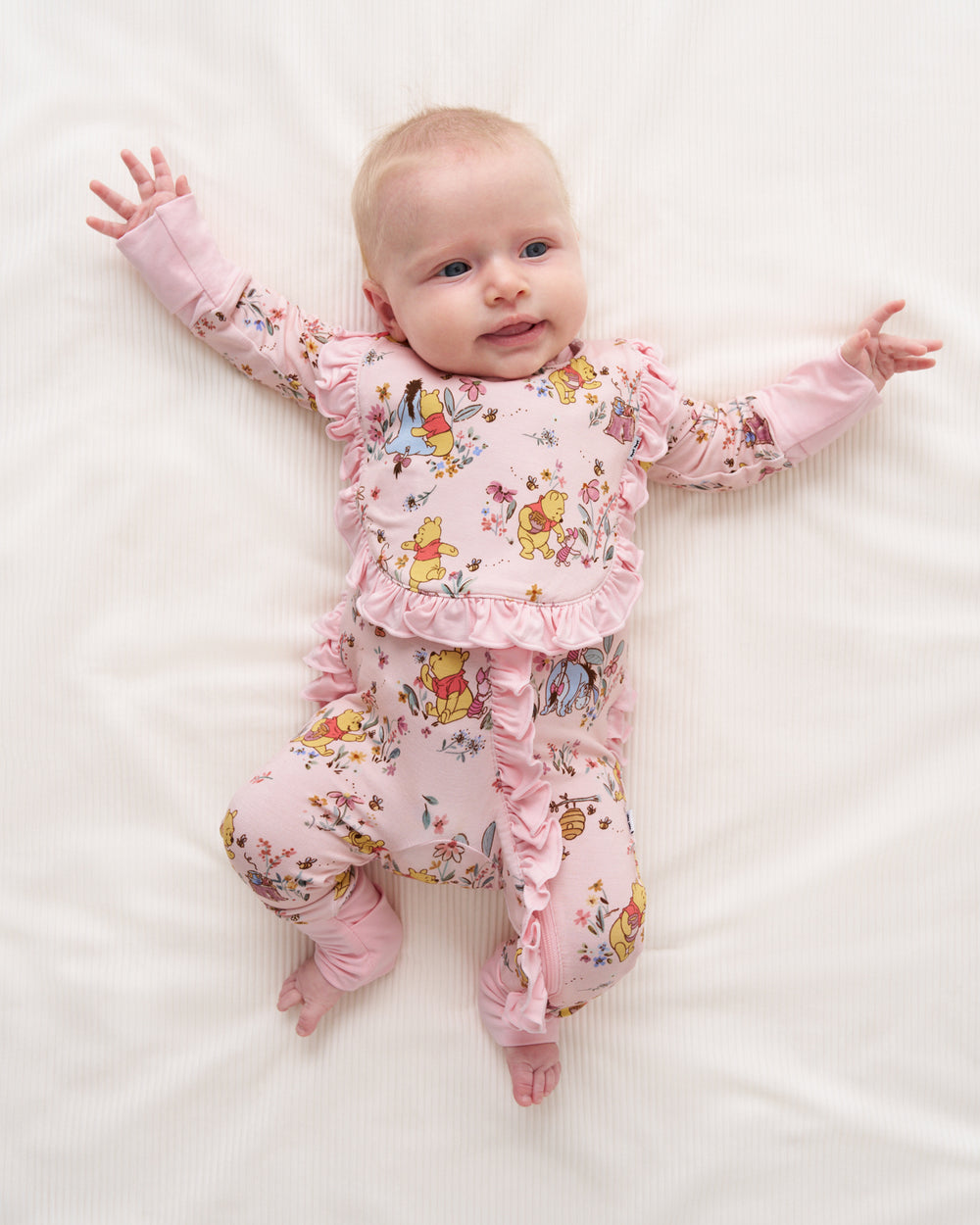 A baby lying on a white textured blanket, wearing a pink long-sleeve footed sleeper with ruffle trim and a colorful cartoon animal print.
