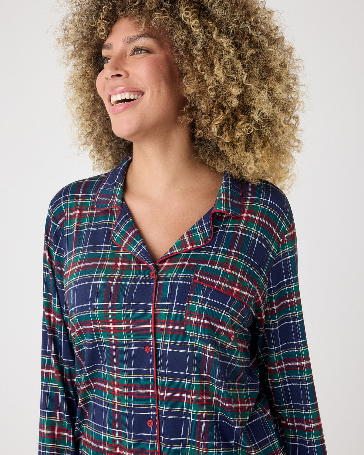 A smiling woman wearing a long-sleeve plaid pajama shirt in navy blue with green, red, and white stripes, photographed against a light gray studio background.