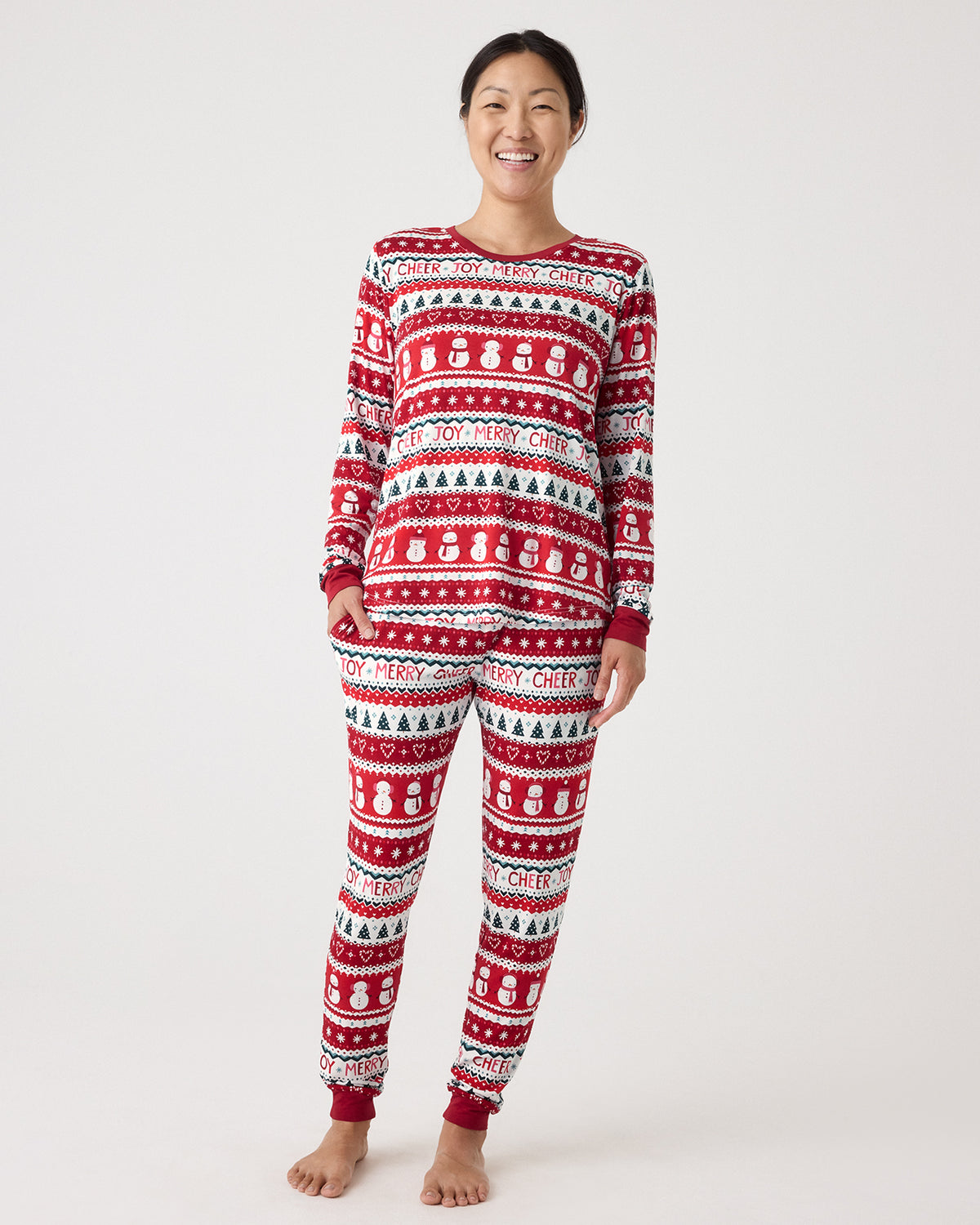 A woman modeling a matching red, white, and green holiday-themed long-sleeve pajama set with snowman and Christmas tree patterns, standing against a light gray studio background.