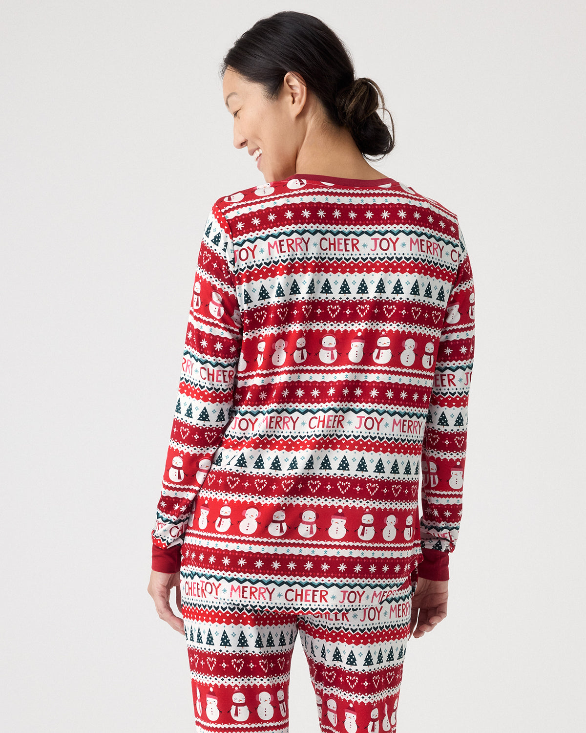 A woman shown from the back wearing a red, white, and green holiday-themed long-sleeve pajama set with a repeating pattern of Christmas cheer quotes, snowballs, and pine trees against a light gray studio background.