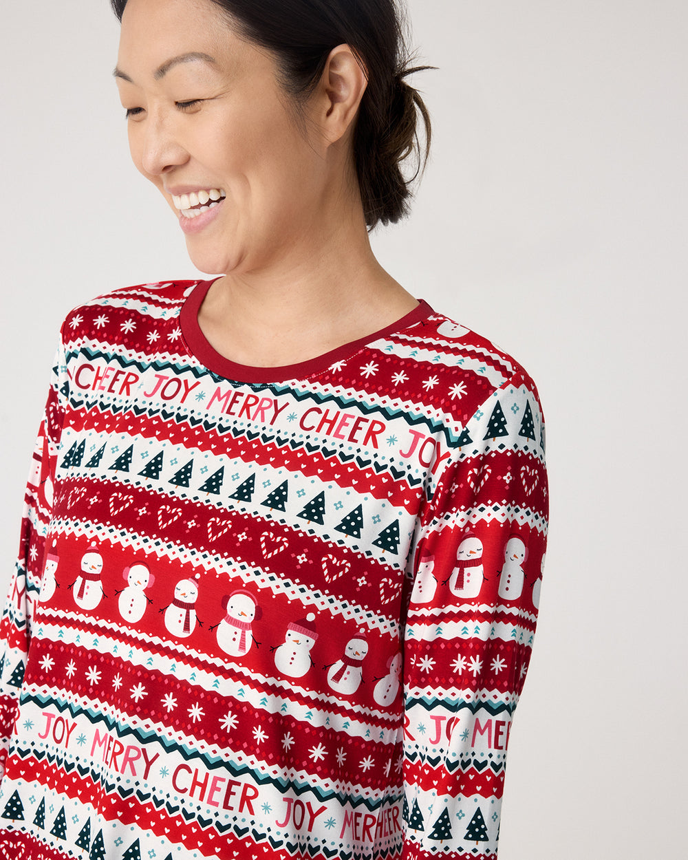 A woman wearing a festive red, white, and navy Christmas sweater with snowman and tree patterns, shown in a close-up shot against a light gray background.