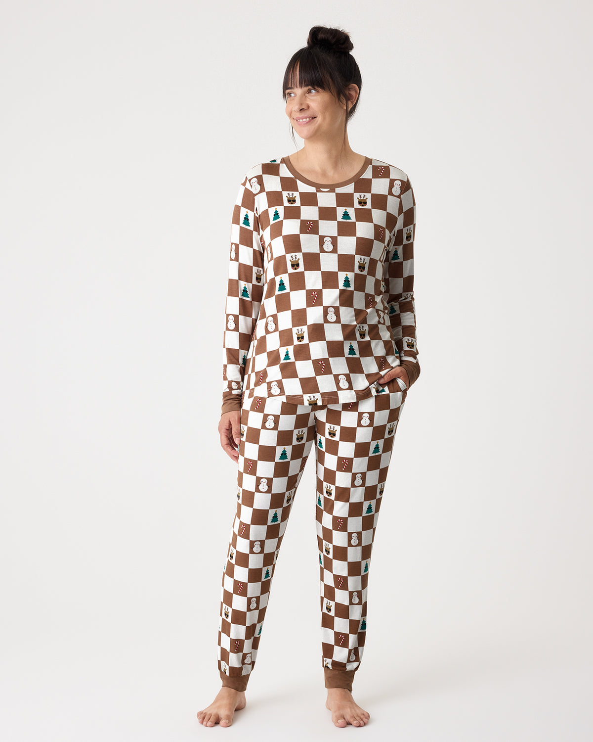 A woman modeling a matching long-sleeve pajama set with a brown and white checkerboard pattern and small holiday-themed icons, standing against a plain white studio background.