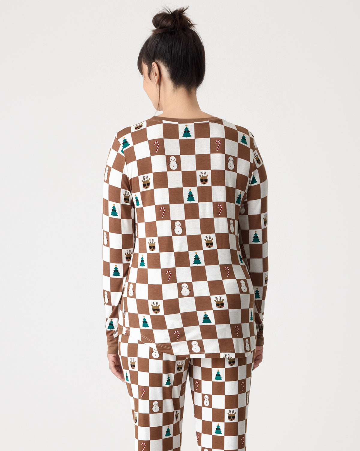 A woman viewed from behind wearing a matching long-sleeve top and pants set with a brown and white checkerboard pattern decorated with small holiday motifs like snowmen, Christmas trees, and reindeer, against a light gray studio background.